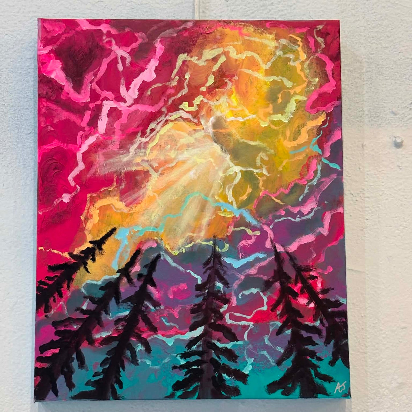 A colorful abstract landscape painting on canvas, featuring black pine tree silhouettes at the bottom against a vibrant background of pink, yellow, orange, purple, and teal. The painting is mounted on a white gallery wall with a small artist label card below it.