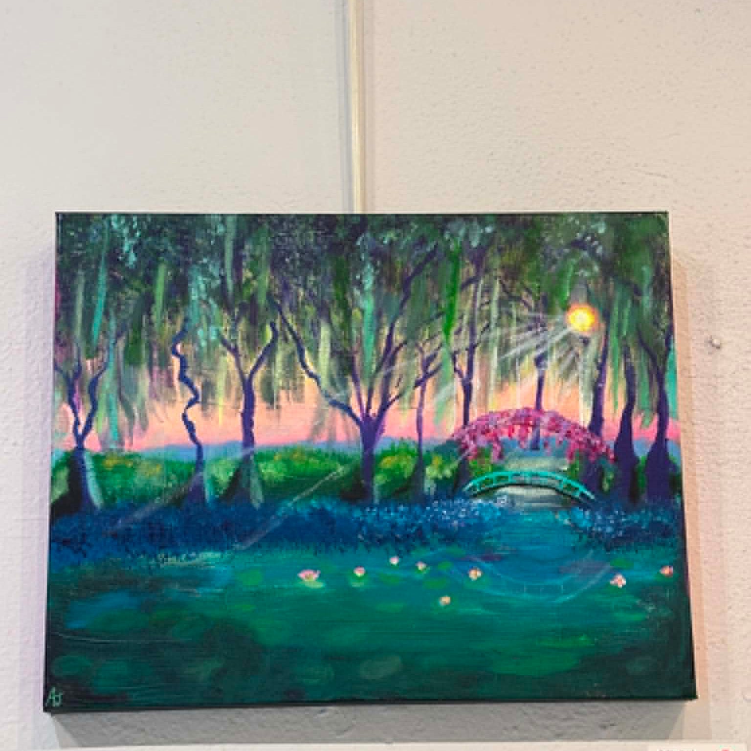 A square canvas painting of a serene pond landscape at dusk, featuring dark green and teal trees, a turquoise arched bridge with pink wisteria hanging overhead, a glowing yellow sun, and small white lotus flowers on the water, displayed on a white wall.